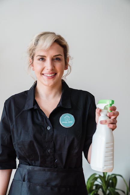 A smiling woman with blonde hair styled in a loose updo, dressed in a black button-up shirt, stands in a room with a plain light grey wall. She is holding a white spray bottle with a green nozzles, de