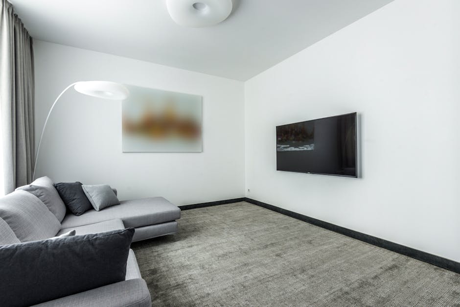 A modern living room with white walls and a large flat-screen television mounted on the left wall. The room features a light gray fabric sofa with two cushions, one black and one gray, positioned near